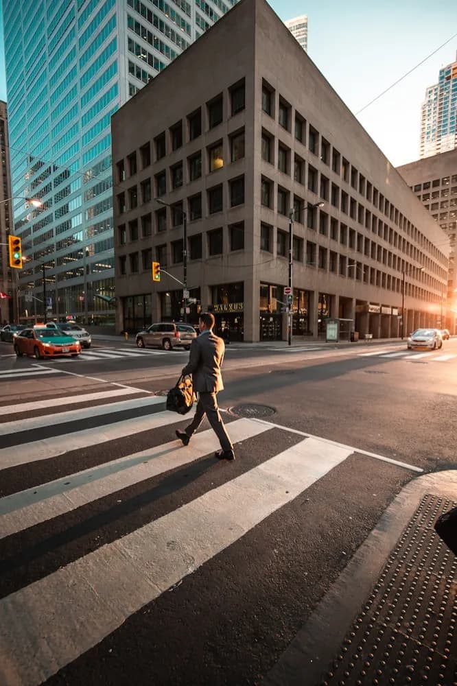 A person crossing a wide street in a financial district at sunrise.