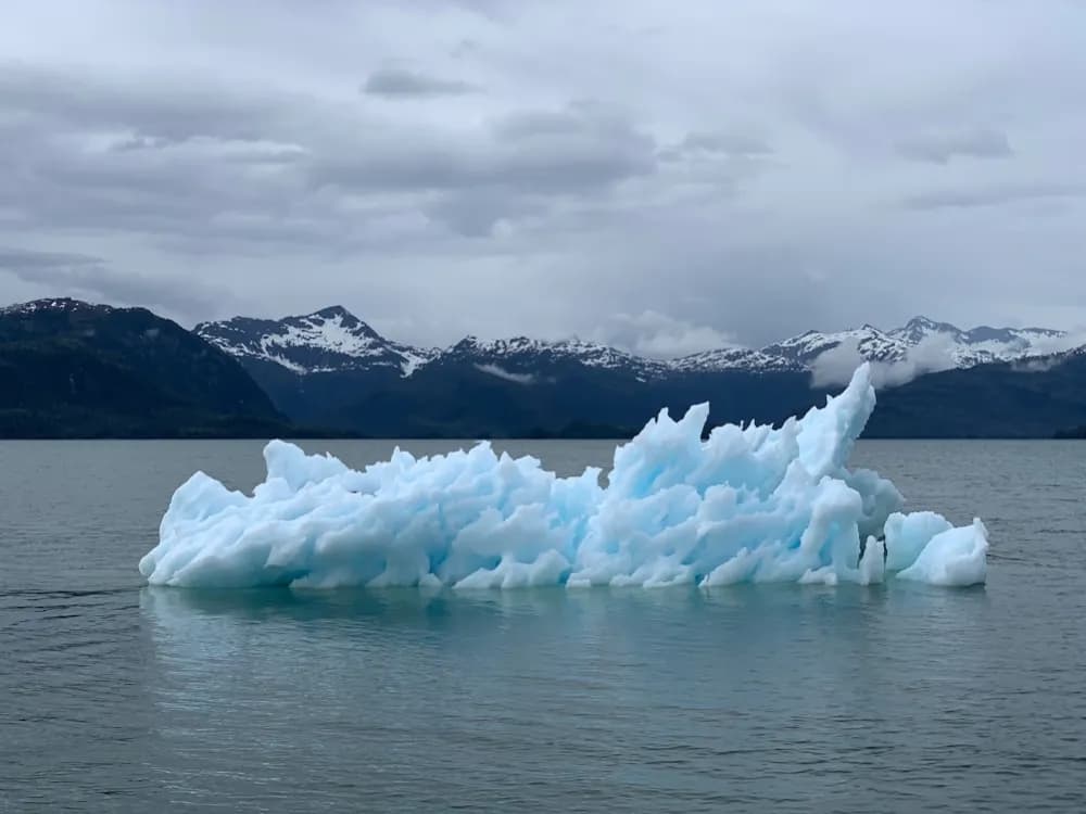 An iceberg on still water in front of snow-covered mountains.