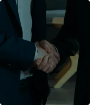 Two people in suits shake hands indoors.